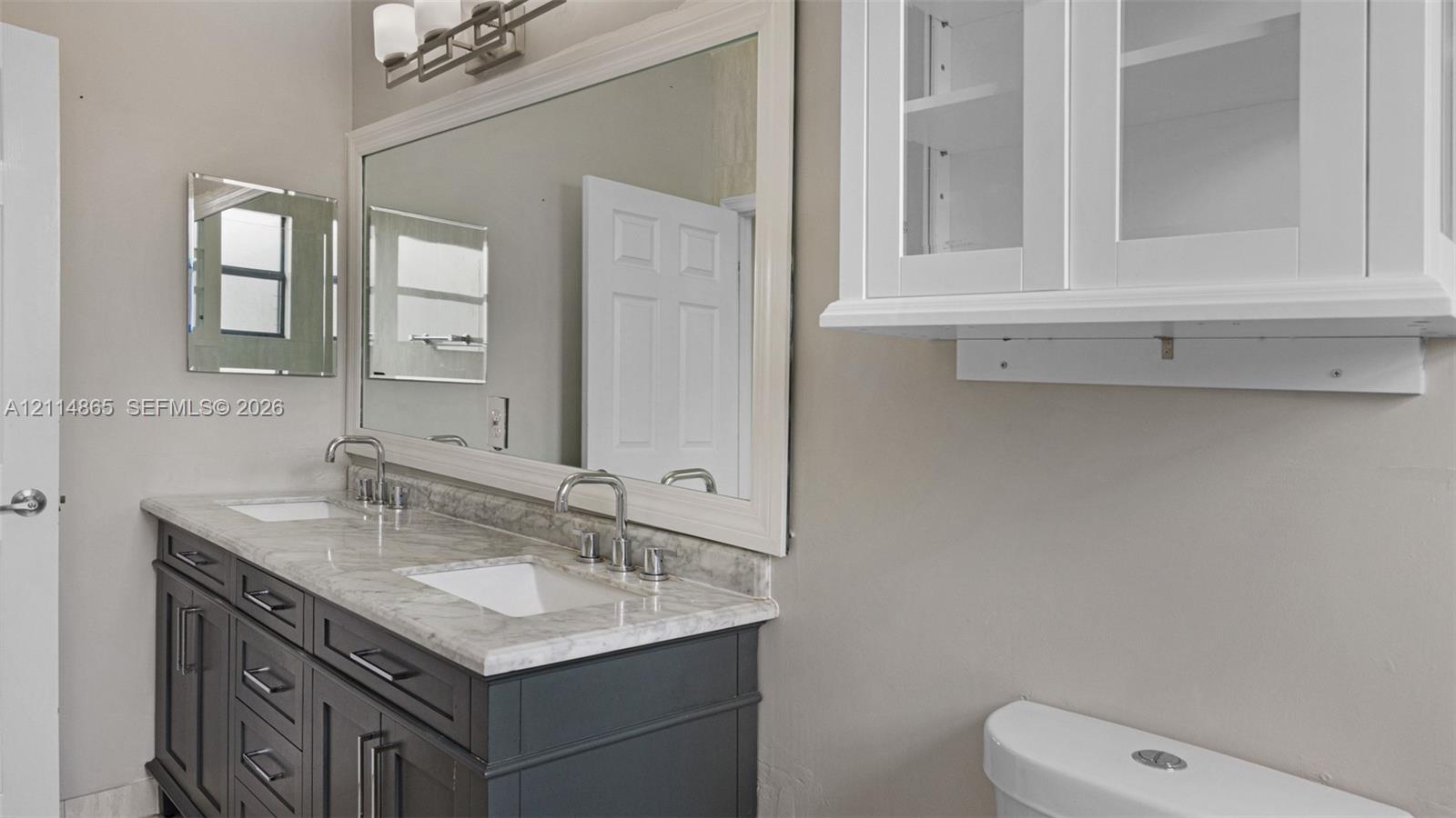 Dual-sink vanity with stone-finish countertop, undermount basins, wall-mounted light fixture, large framed mirror, and white built-in upper cabinetry with glass-front doors