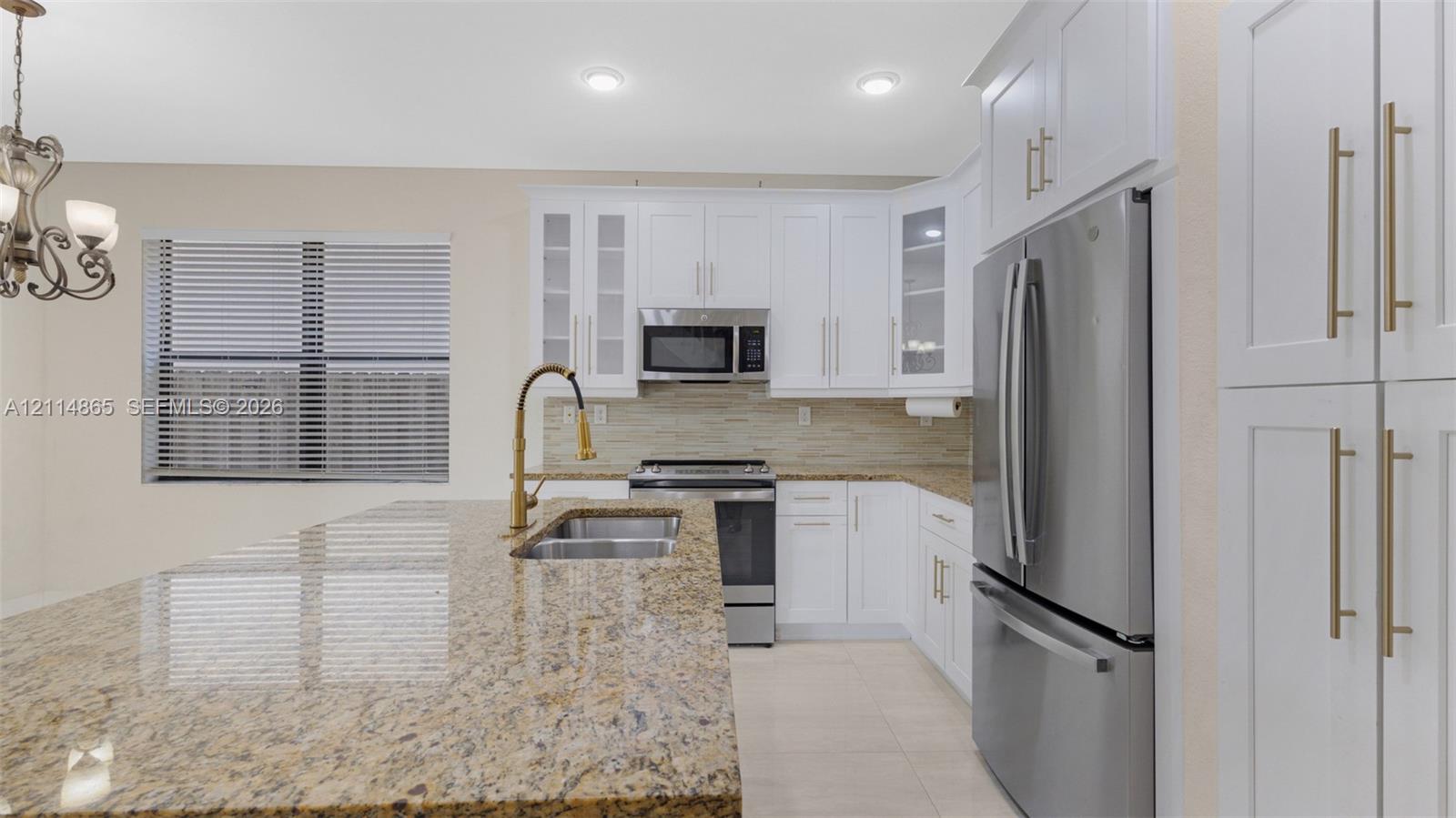 Kitchen featuring white shaker-style cabinetry, stone countertops, a central island with an integrated double-basin sink and arched faucet, stainless steel appliances, and a tile backsplash