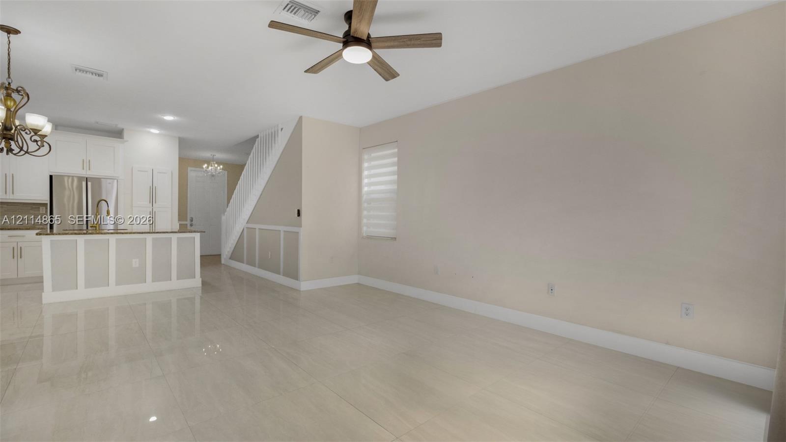 Open living area featuring large-format polished tile flooring, neutral wall tones, beadboard wainscoting, multi-blade ceiling fan, and kitchen with white cabinetry and island