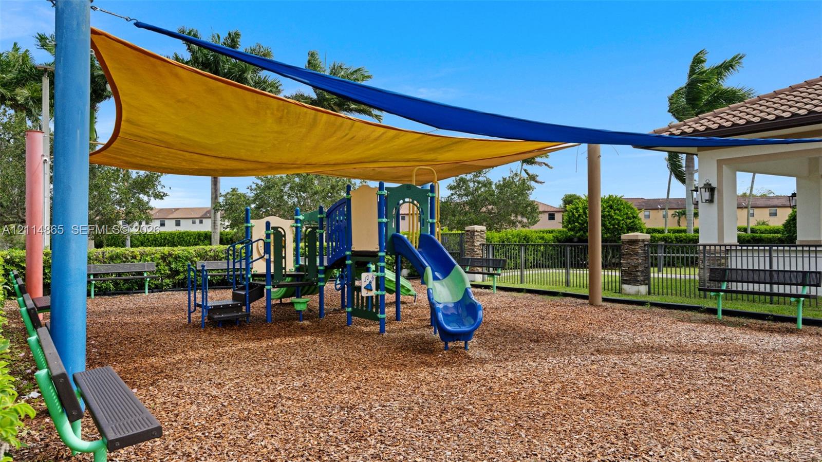 Play area featuring multi-level climbing equipment, a blue slide, structural shade sails, and perimeter metal fencing
