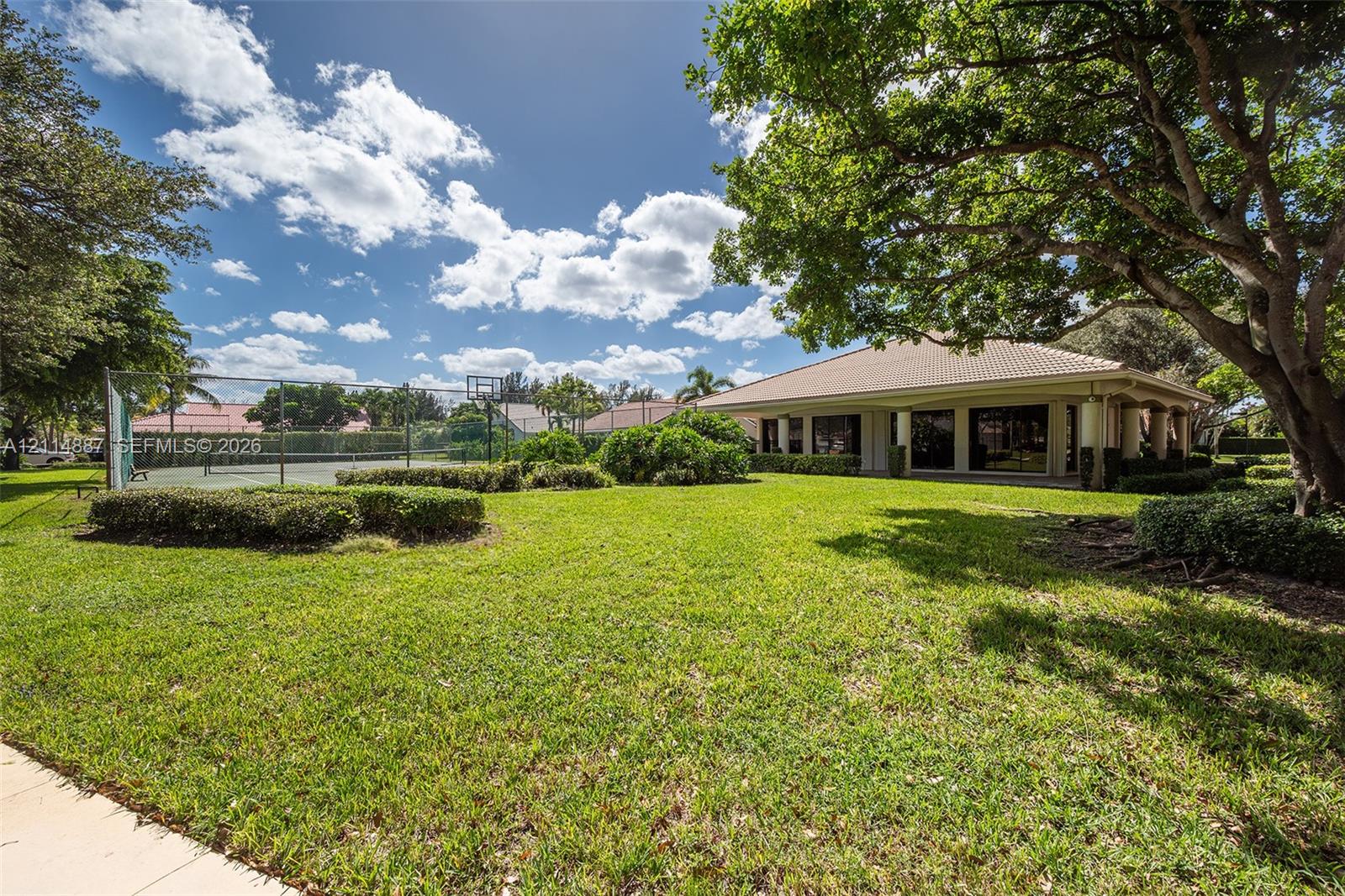 Expansive rear grounds featuring a fenced tennis court, manicured lawn, mature shade trees, and a covered patio