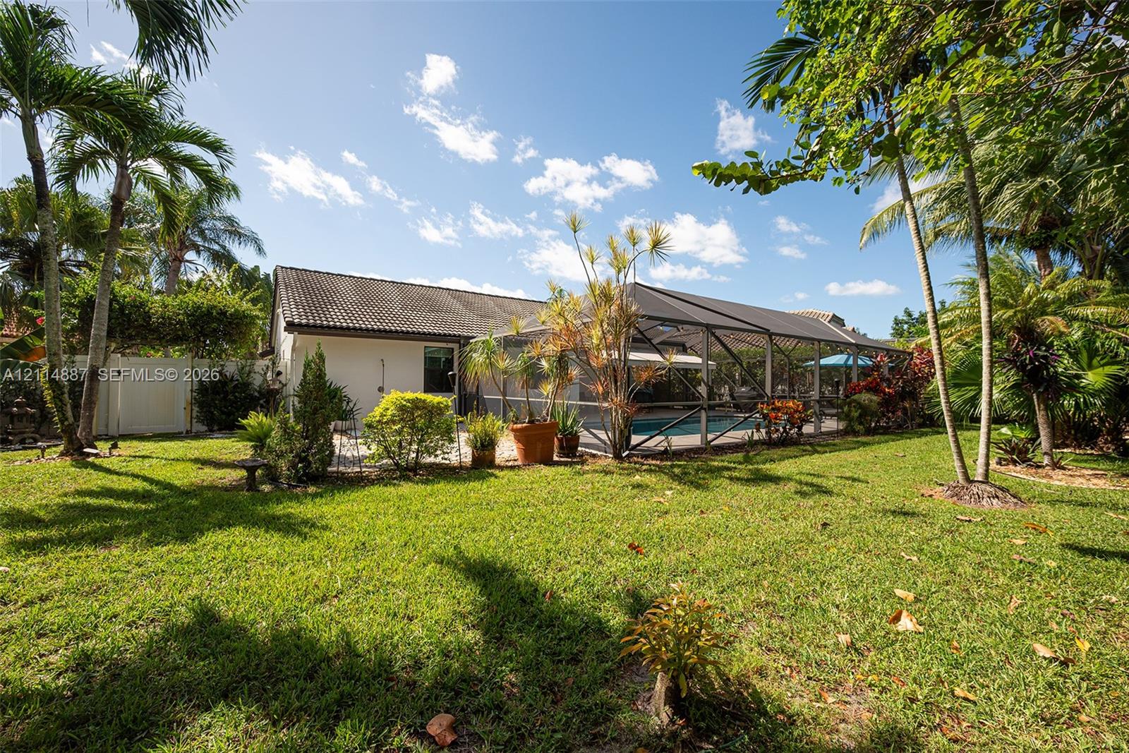 Spacious backyard featuring a manicured lawn, perimeter vinyl fencing, mature palm trees, and an enclosed screened-in pool area