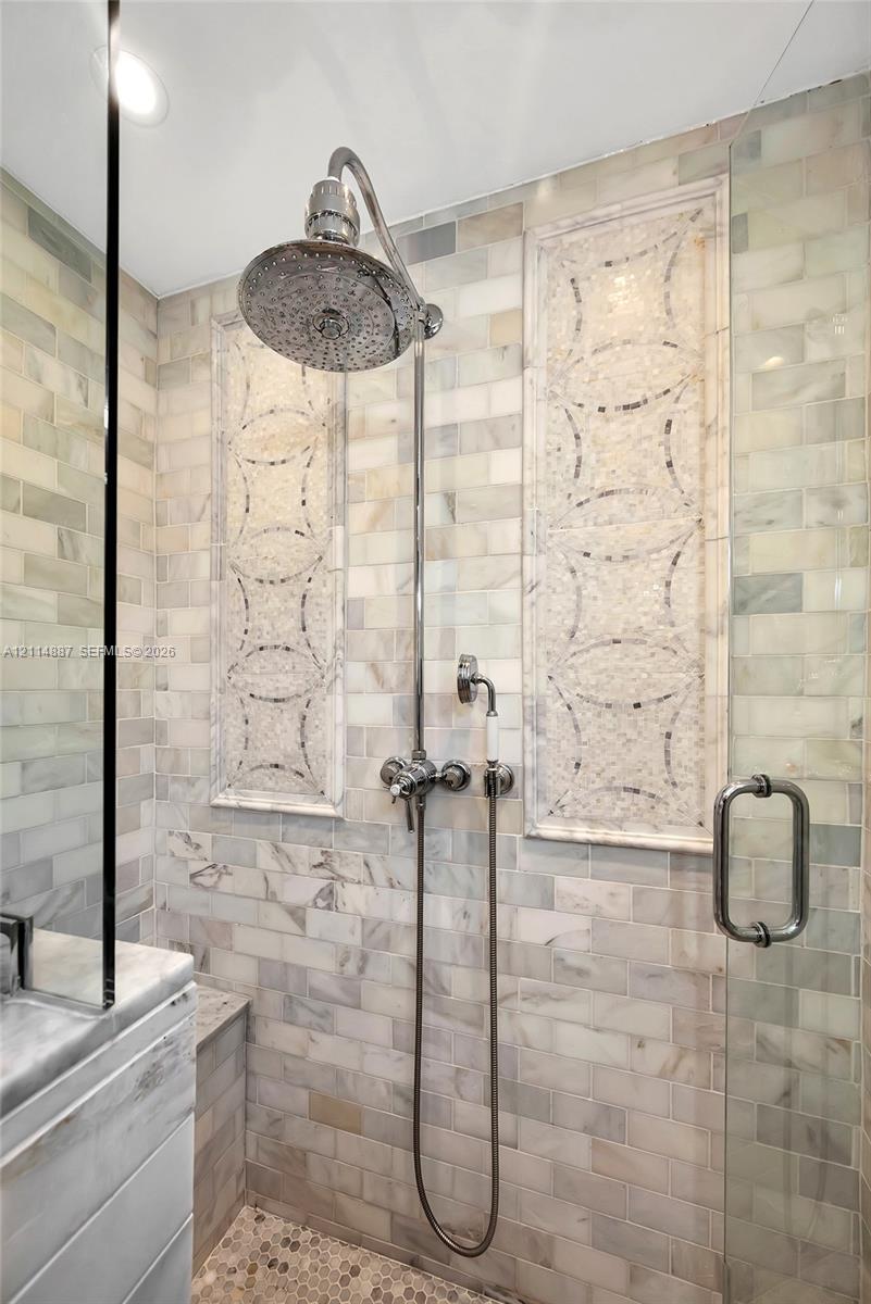 Walk-in shower featuring brick-pattern stone wall tile, decorative inset mosaic panels, pebble flooring, polished metal rainfall showerhead with handheld attachment, and a glass door enclosure