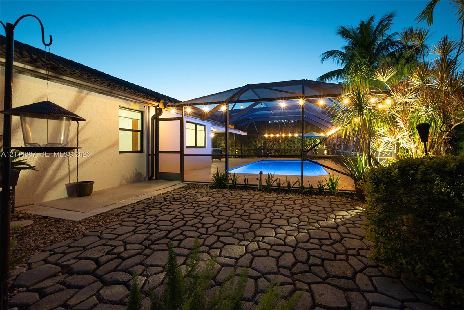 Screened-in outdoor pool area with stone-patterned paver patio, stucco exterior wall, and lush perimeter landscaping