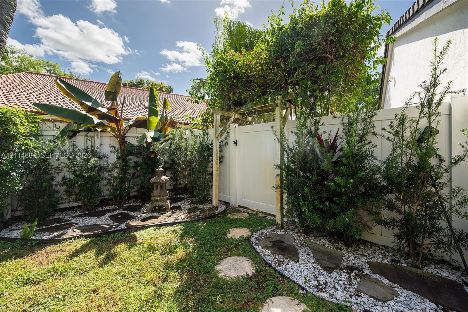 Enclosed backyard featuring white vinyl fencing, a decorative wooden arbor, stone paver pathways, and landscape plantings