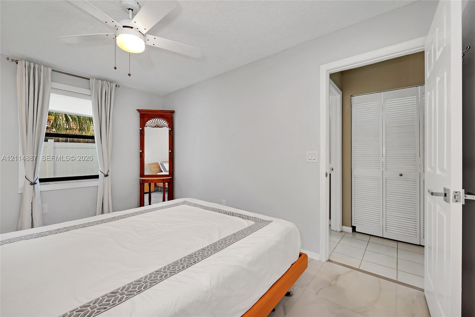Neutral-toned interior featuring large-format tile flooring, white bi-fold closet doors, and a ceiling-mounted light fixture with a multi-blade fan