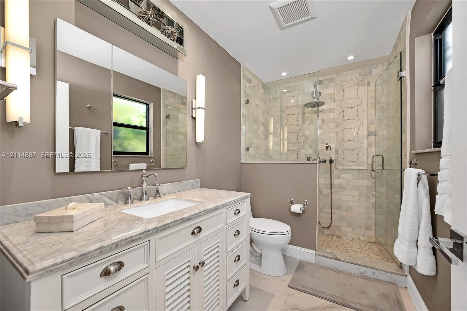 White vanity with stone countertop and undermount sink, walk-in shower with stone tile surround and decorative inlay, frameless glass enclosure, dual vertical wall sconces, and rectangular wall mirror