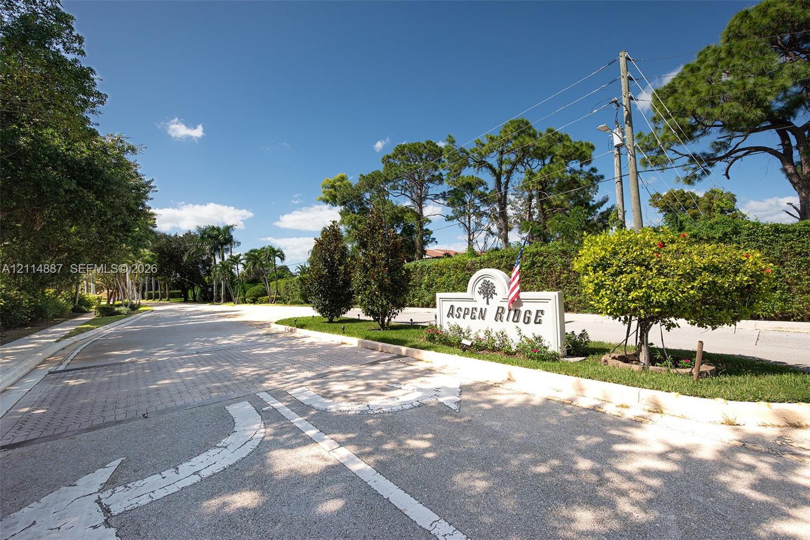 Community entrance featuring stone-paver roadway, landscaped grounds, mature pine and palm trees, and a monument sign