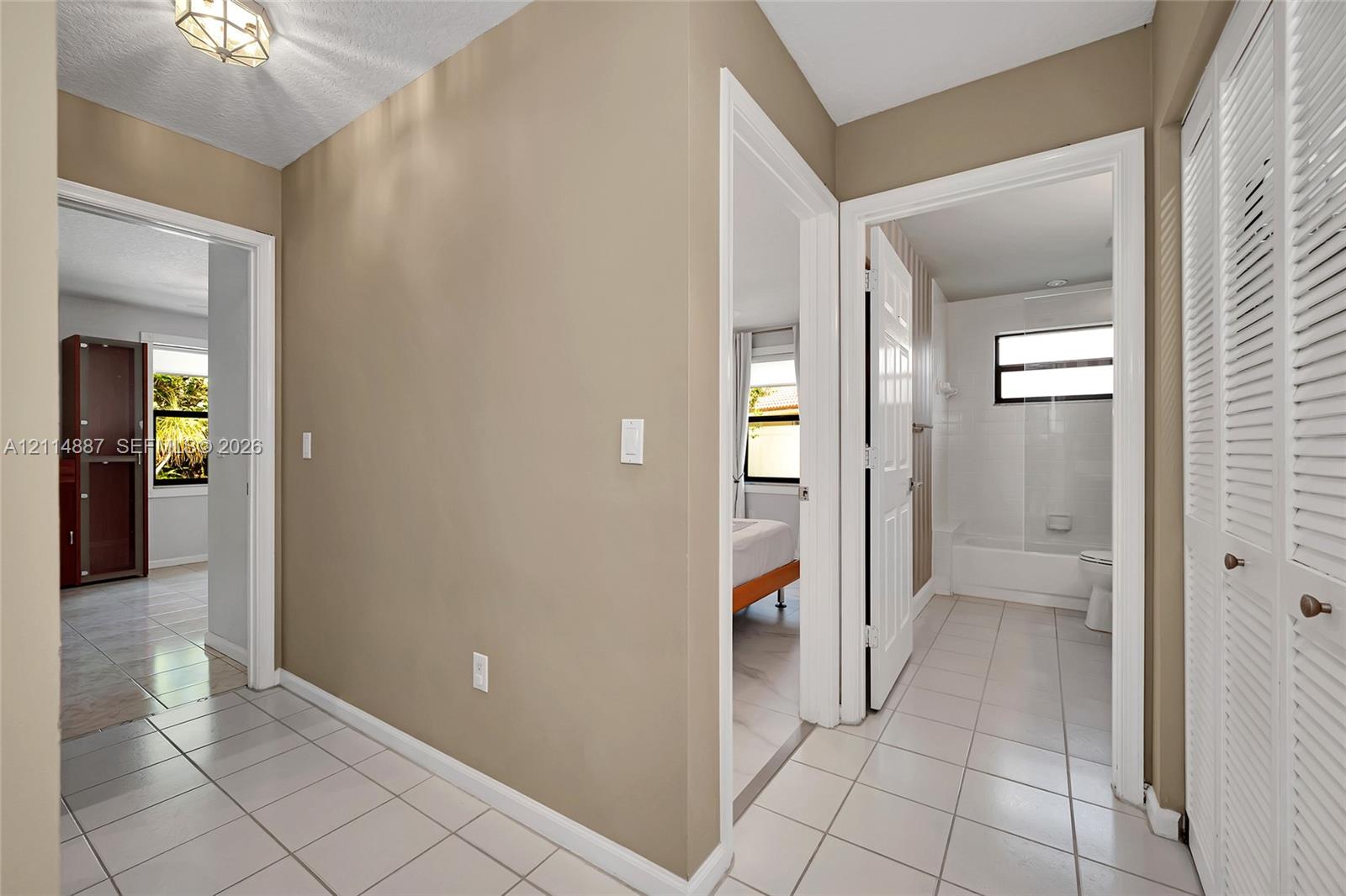 Neutral-toned hallway featuring ceramic tile flooring, recessed geometric lighting fixture, and floor-to-ceiling louvered closet doors