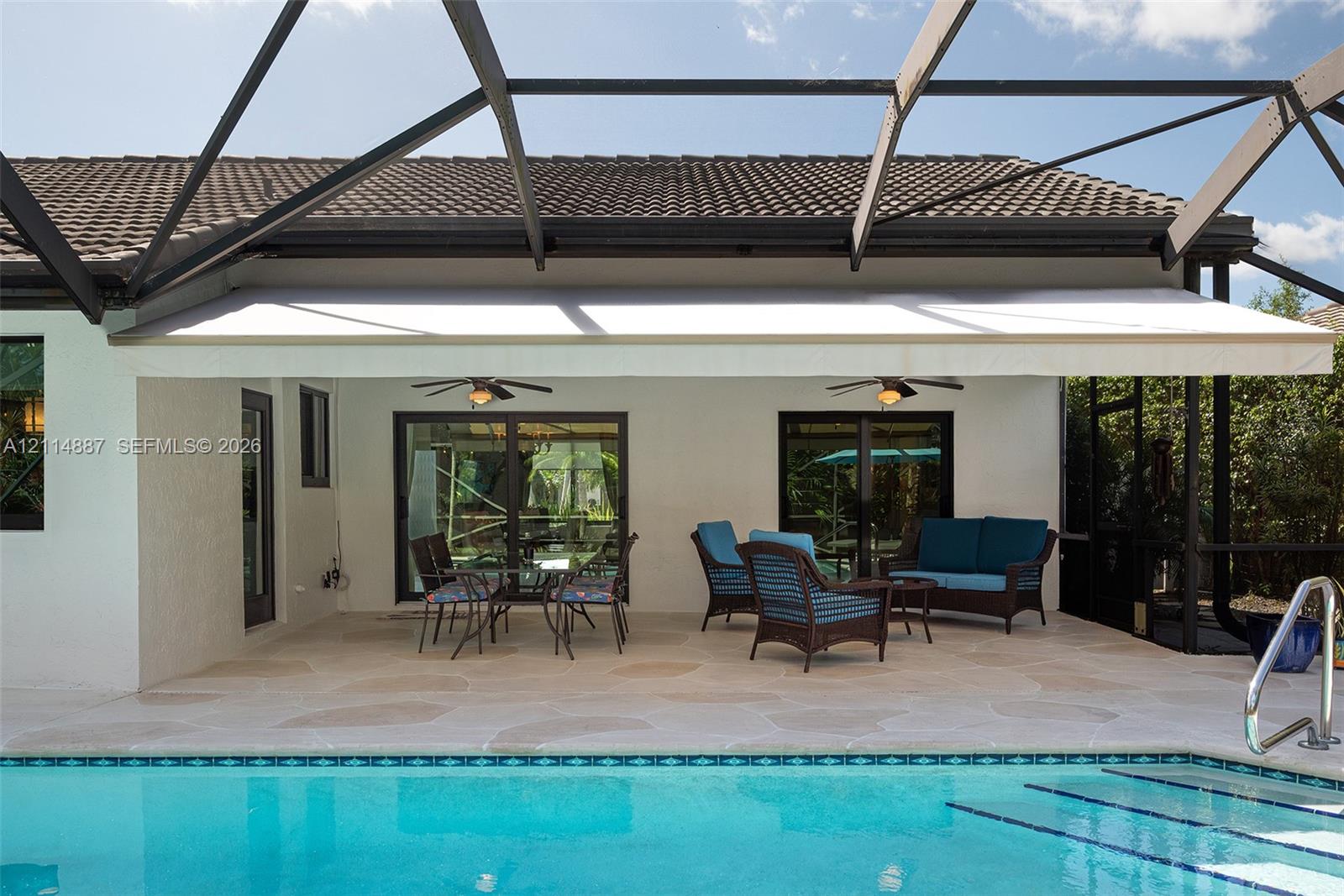 Covered patio featuring natural stone pavers, a retractable awning, dual ceiling fans, and a screened enclosure overlooking a swimming pool