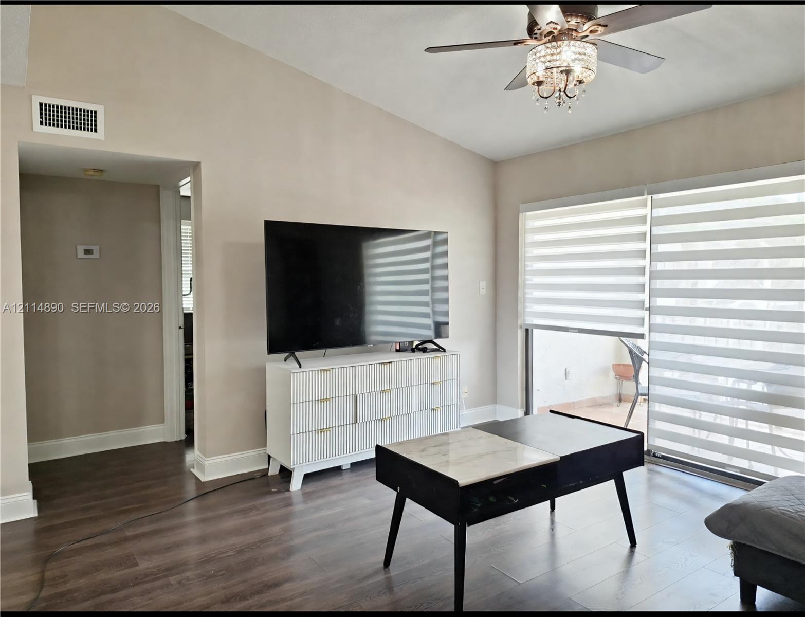 Vaulted ceiling featuring a central ceiling fan with integrated crystal chandelier, wood-finish flooring, and sliding glass doors with roller shades
