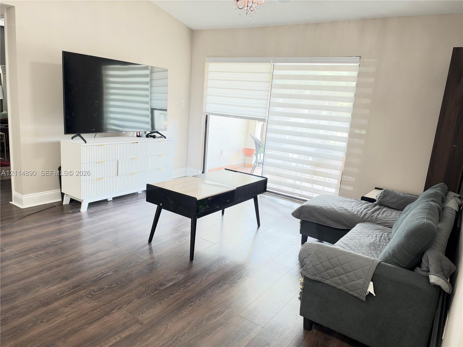 Vaulted ceiling with a central pendant light fixture, dark wood-finish flooring, and large sliding glass doors featuring horizontal banded shades