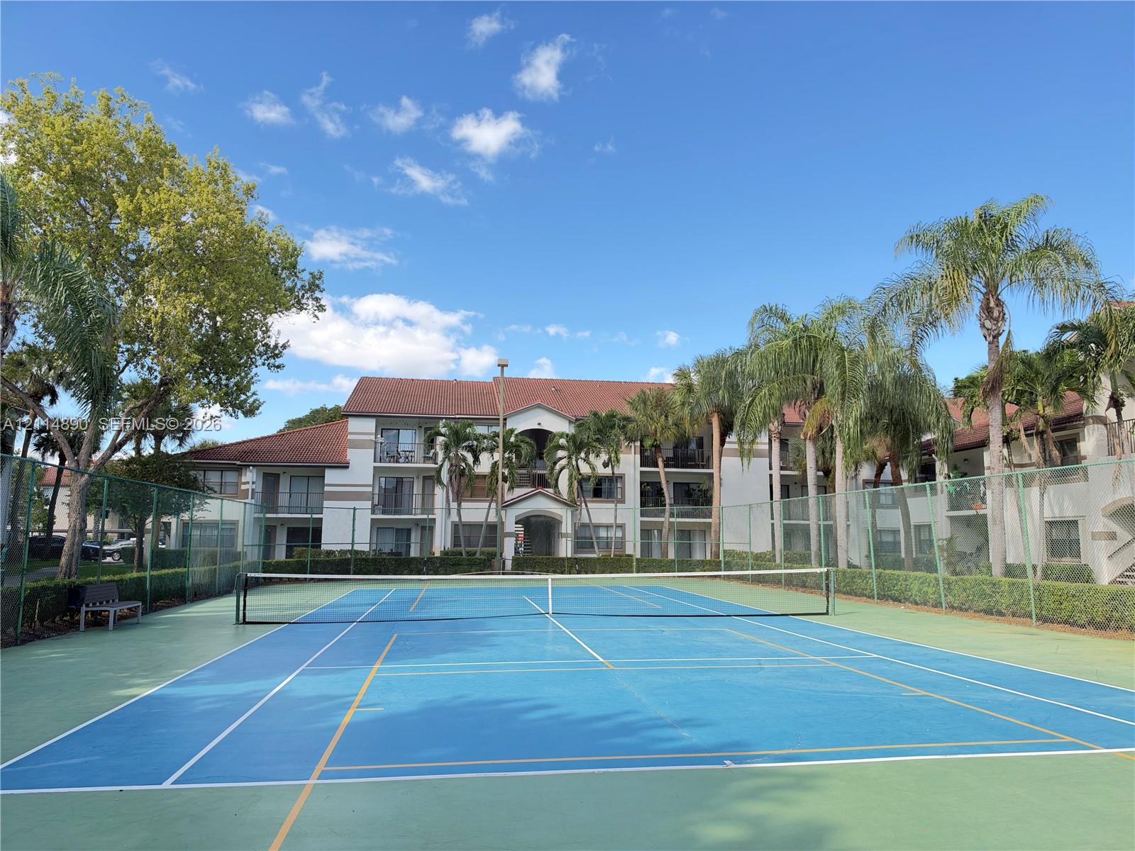 Hard-surface tennis court enclosed by perimeter fencing and landscaped borders, featuring multi-story residential architecture with terracotta-style roof tiling