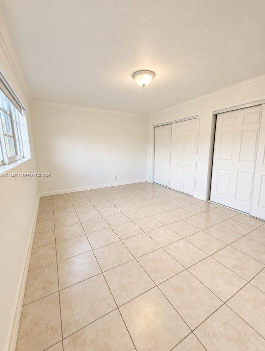 Room featuring neutral tile flooring, crown molding, multi-pane window, and dual sliding-panel closets with six-panel doors Room featuring neutral tile flooring, crown molding, multi-pane window, and dual sliding-panel closets with six-panel doors