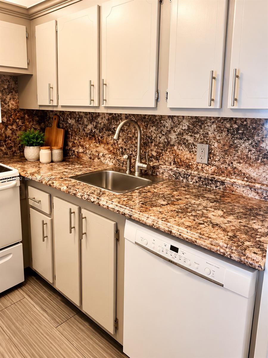Kitchen featuring white cabinetry with brushed metal hardware, stone-patterned countertops, matching full-height stone-patterned backsplash, stainless steel sink with high-arc faucet, and wood-finish tile flooring