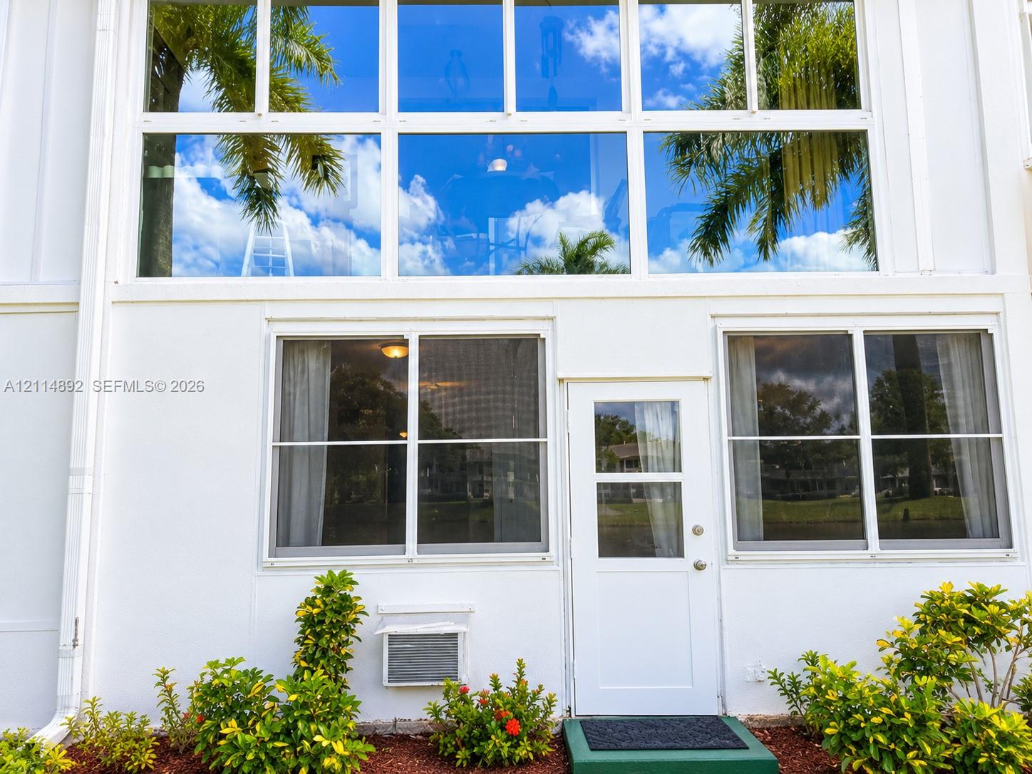 Exterior facade featuring multi-pane floor-to-ceiling windows, white siding, and a pedestrian door with a glass insert, complemented by foundation landscaping