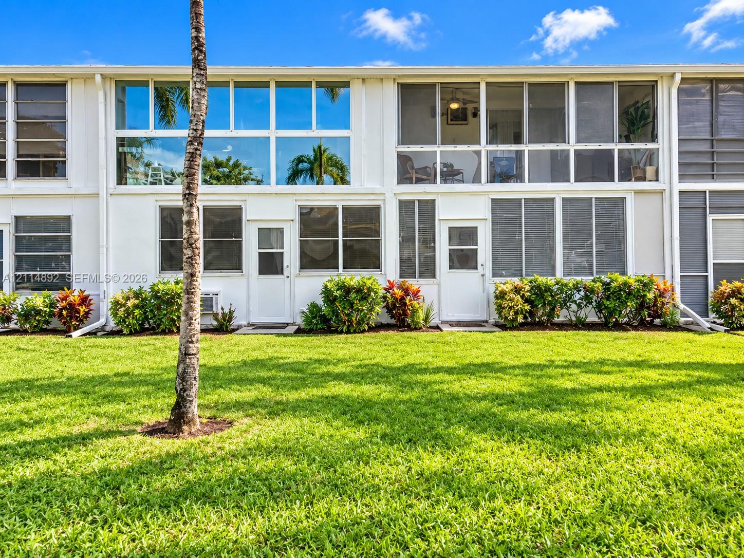 Multi-level building exterior featuring white stucco facade, expansive floor-to-ceiling windows, screened-in upper-level enclosures, and manicured green space with mature landscaping