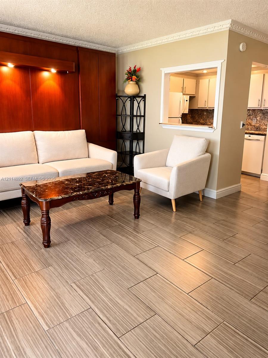 Living area featuring wood-finish tile flooring, decorative crown molding, a wood-paneled accent wall with integrated recessed lighting, and a service pass-through window