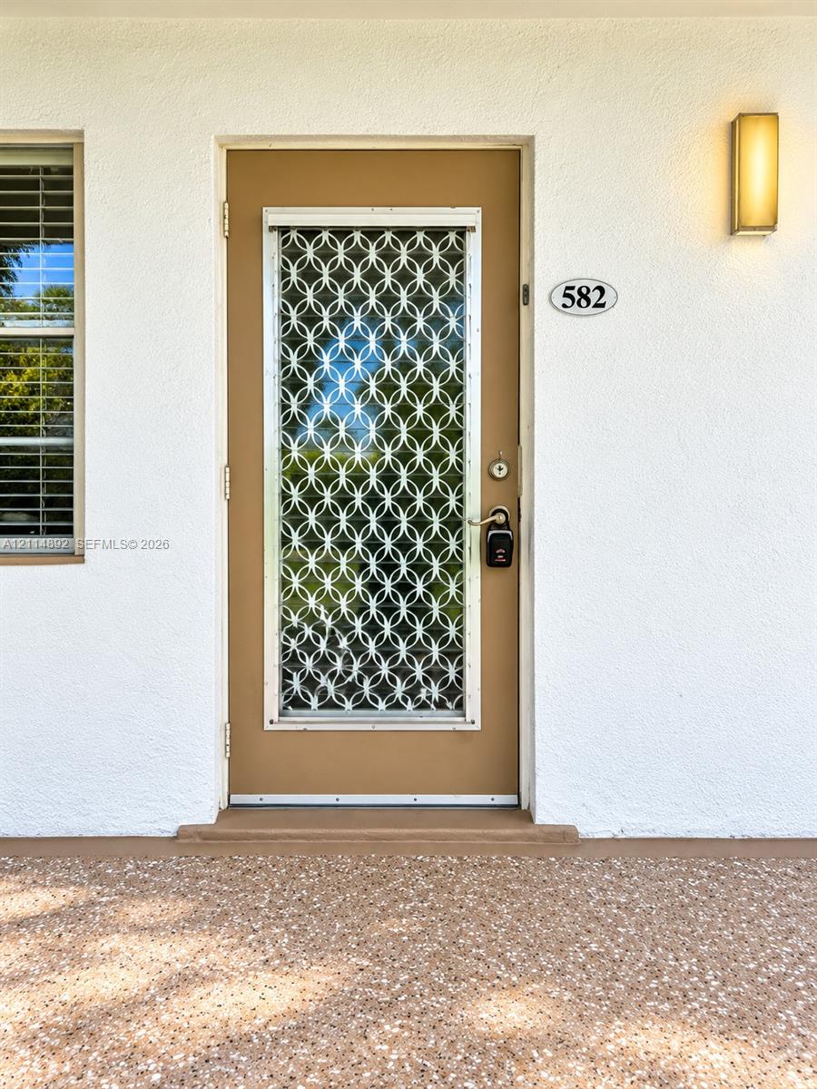 Entrance featuring a solid-color door with an integrated geometric pattern screen, textured stucco exterior wall, rectangular wall-mounted light fixture, and aggregate concrete porch finish