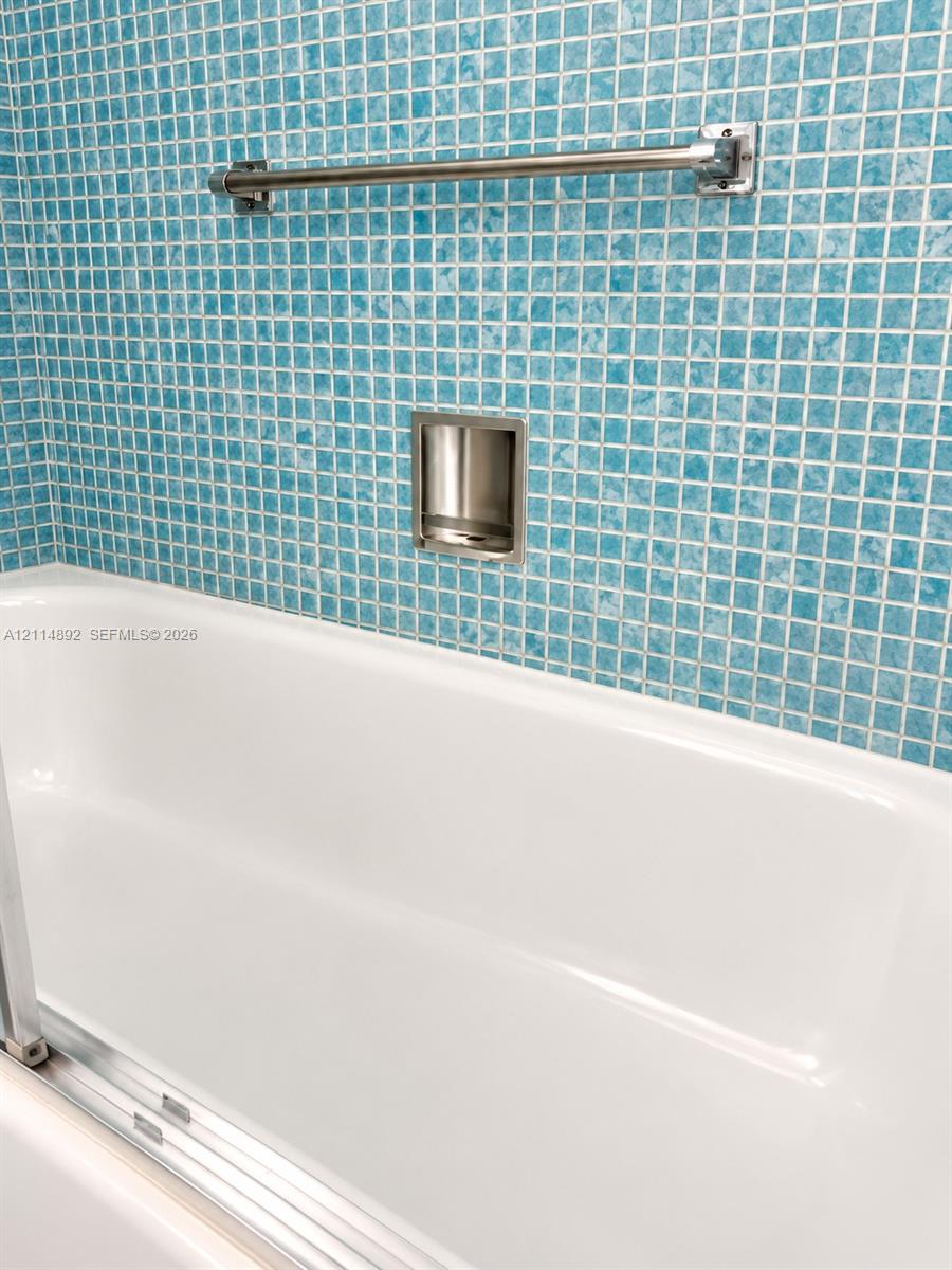White bathtub with integrated glass door track, complemented by blue mosaic tile surround, built-in recessed soap niche, and polished metal towel bar