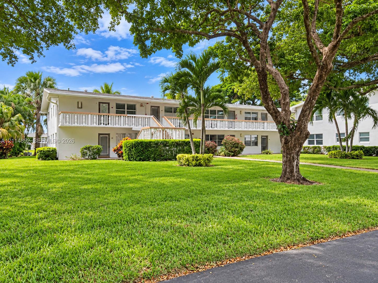 Multi-level exterior featuring a second-story balcony with railings, manicured lawn, established landscaping, and mature trees