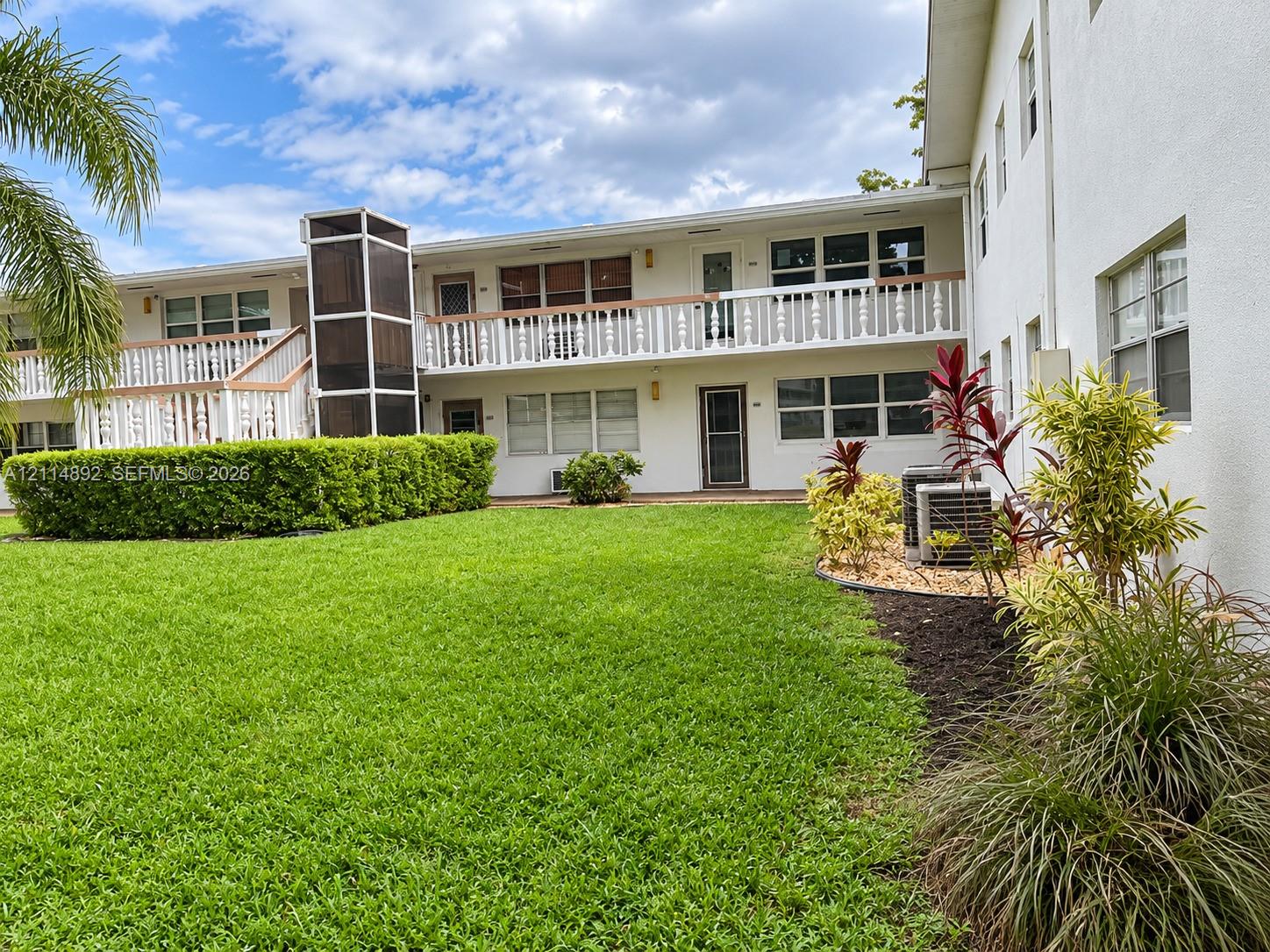 Two-story exterior featuring white stucco siding, a wrap-around balcony with decorative balustrades, an integrated elevator shaft, and landscaped grounds with mature shrubbery
