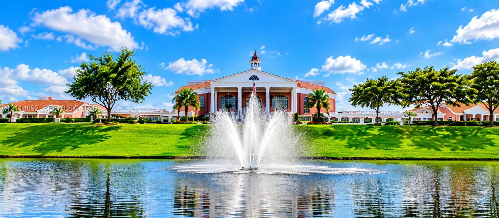 Colonial-inspired architecture featuring a central cupola and multi-columned portico, situated on a manicured waterfront lot with a central water feature and mature landscaping