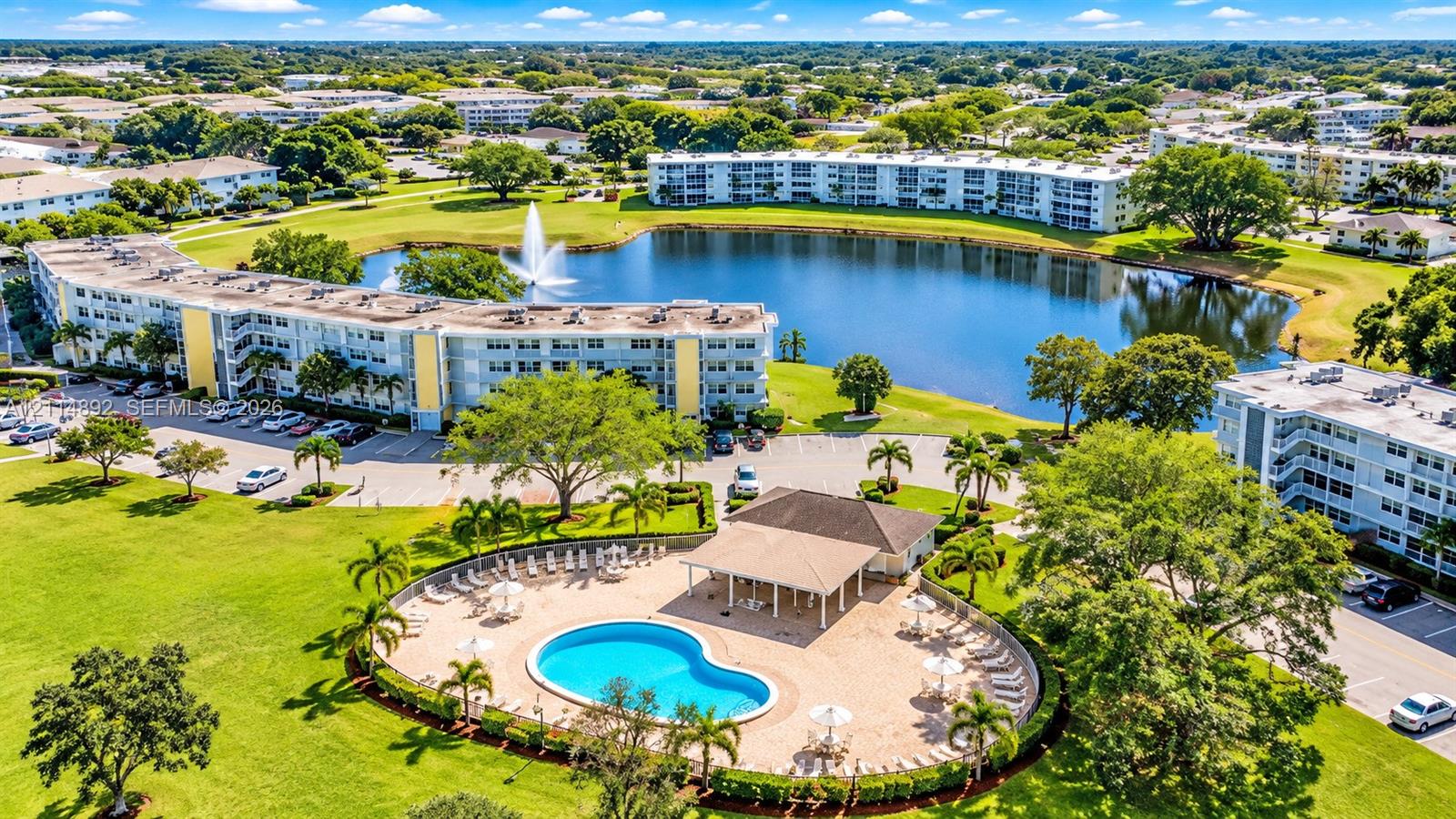 Aerial perspective of multi-unit residential grounds featuring a swimming pool, covered cabana, expansive pond with a central fountain, and mature landscaping