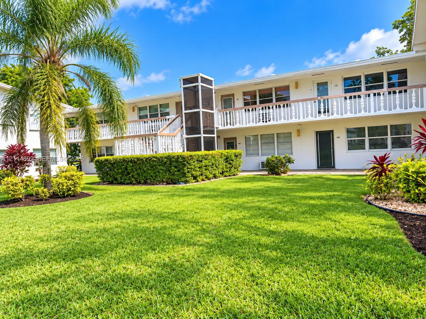 Two-story exterior featuring white stucco siding, a second-level railed balcony, manicured lawn, and mature palm tree landscaping