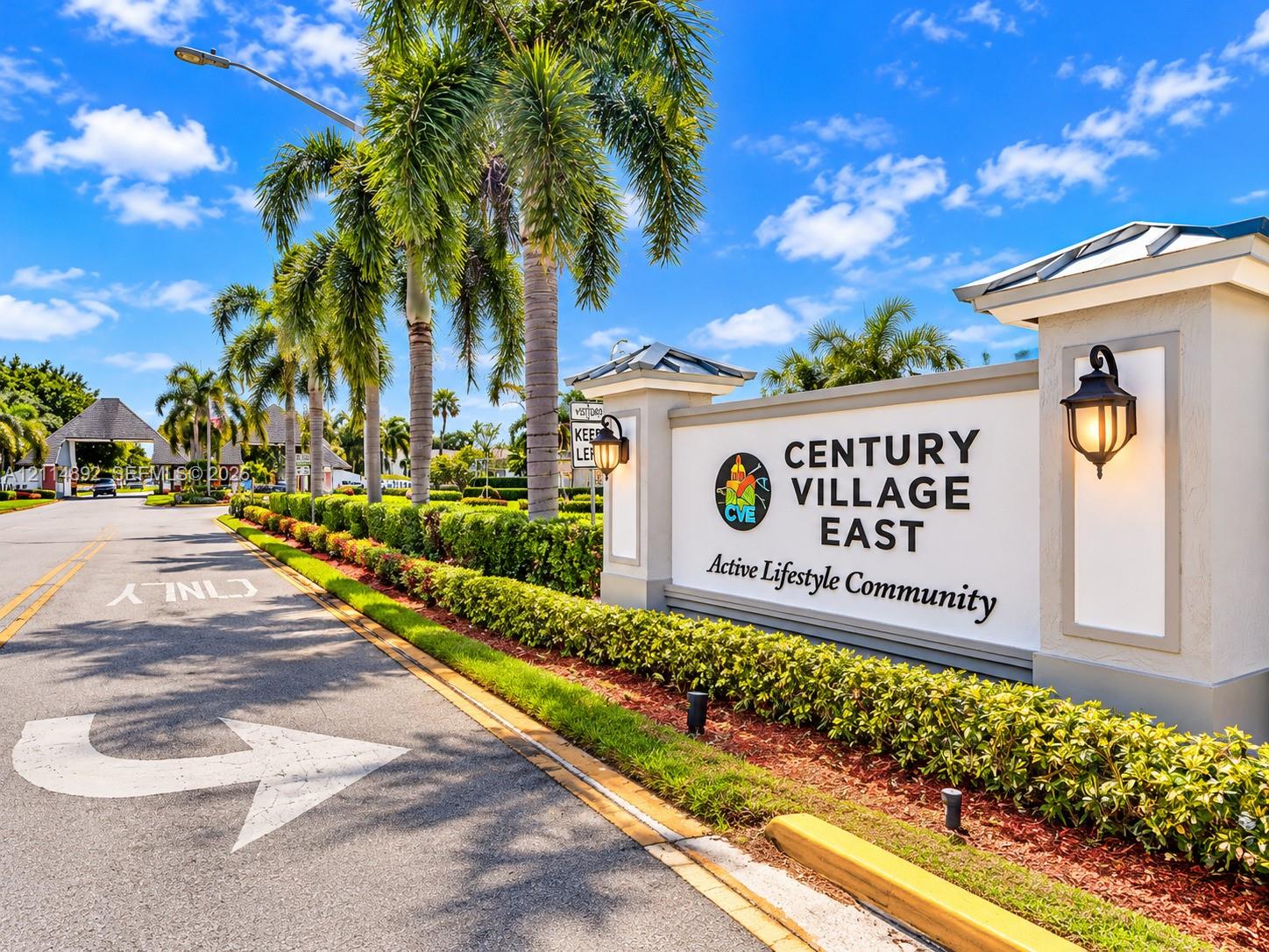 Community entrance featuring manicured hedges, mature palm trees, decorative wall-mounted lighting, and paved roadway access