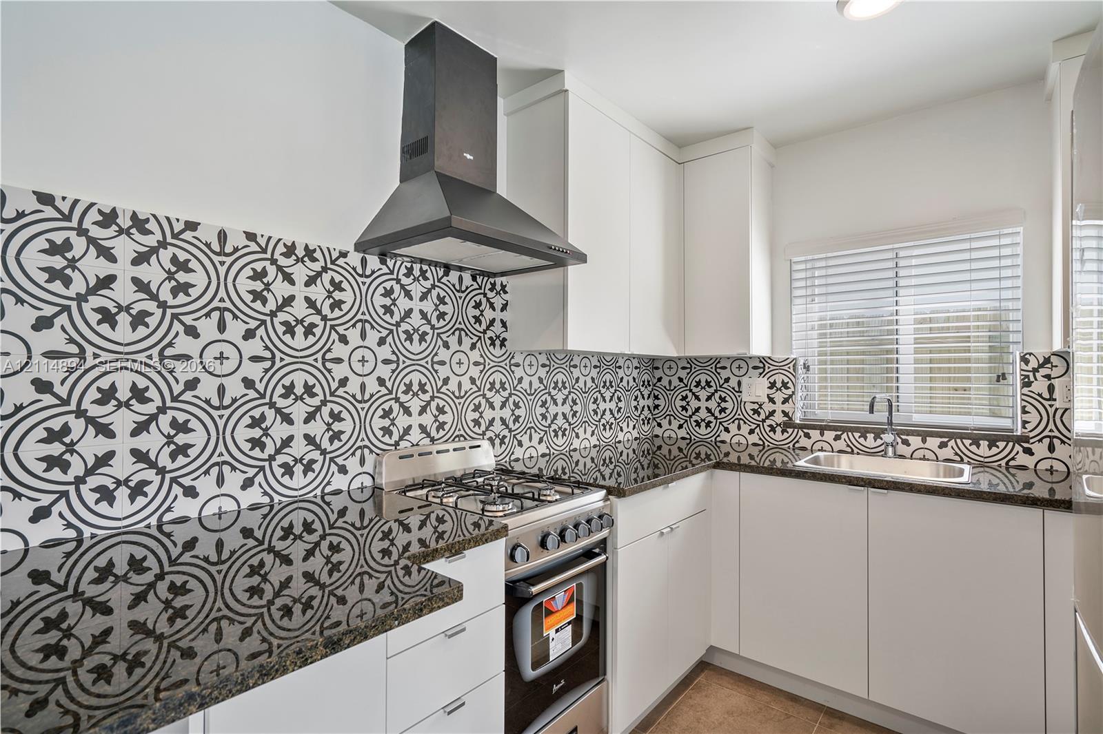 Kitchen featuring white flat-panel cabinetry, dark stone countertops, patterned tile backsplash, stainless steel gas range, and a wall-mounted range hood