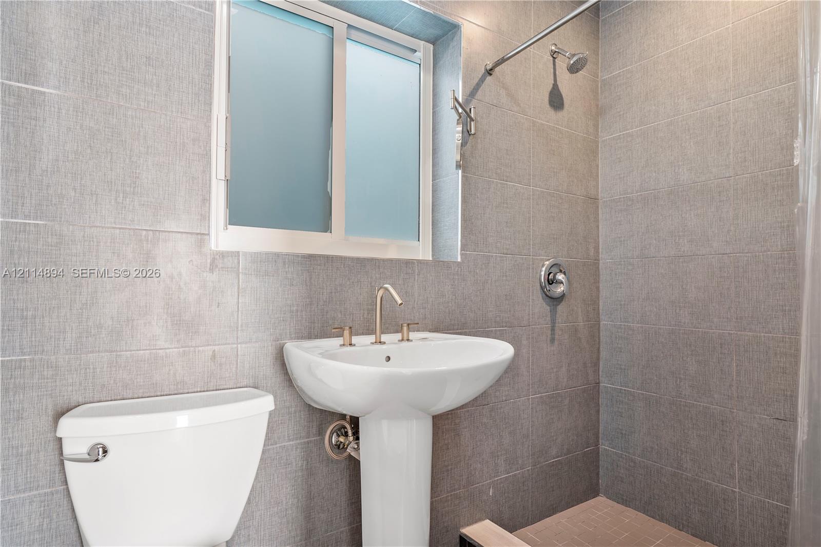 Full-height gray textured wall tiling, white pedestal sink with metal widespread faucet, and shower head with integrated control handle