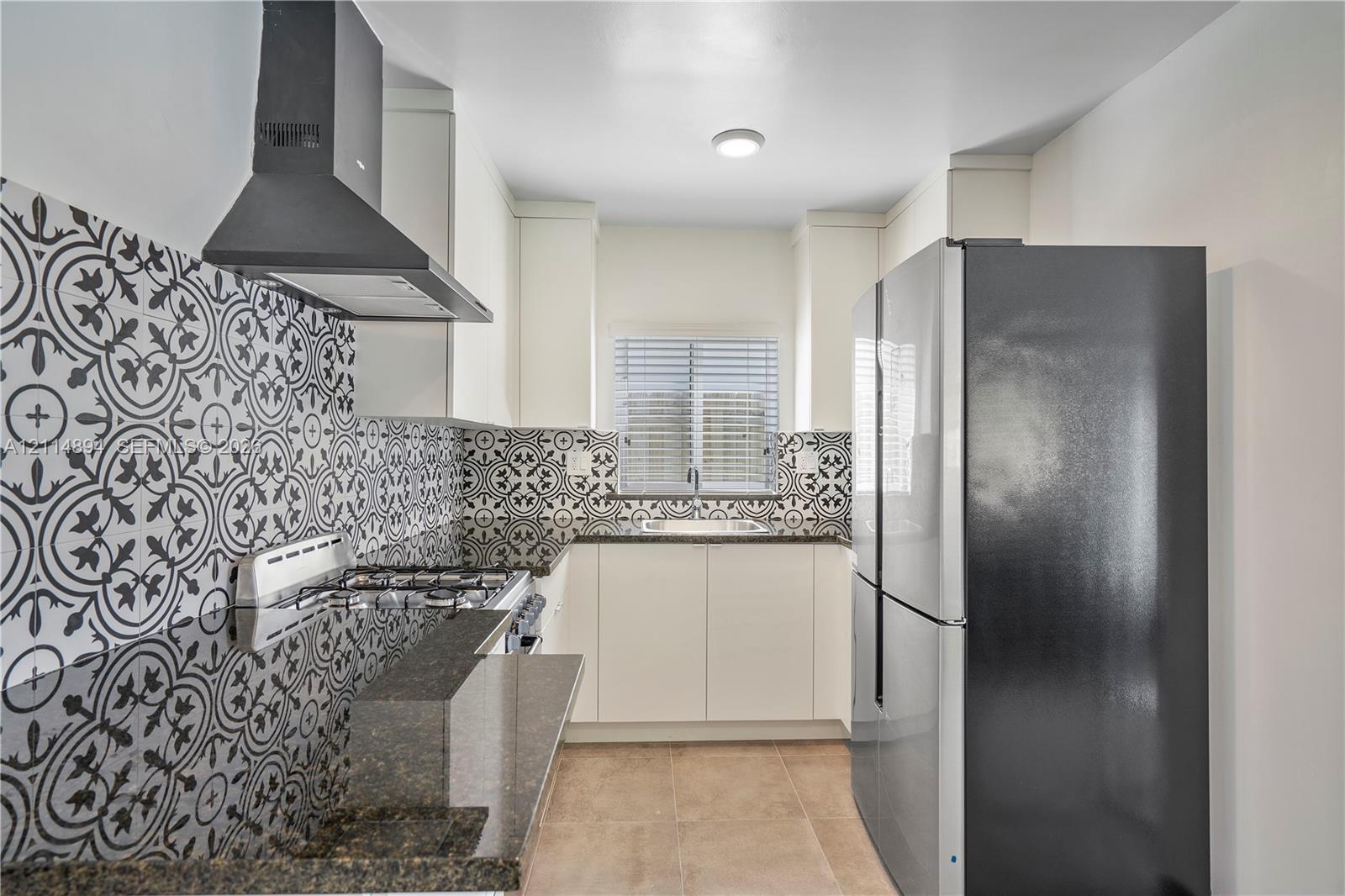 Kitchen featuring patterned tile backsplash, dark stone-finish countertops, matte black range hood, sleek white cabinetry, and stainless steel refrigerator