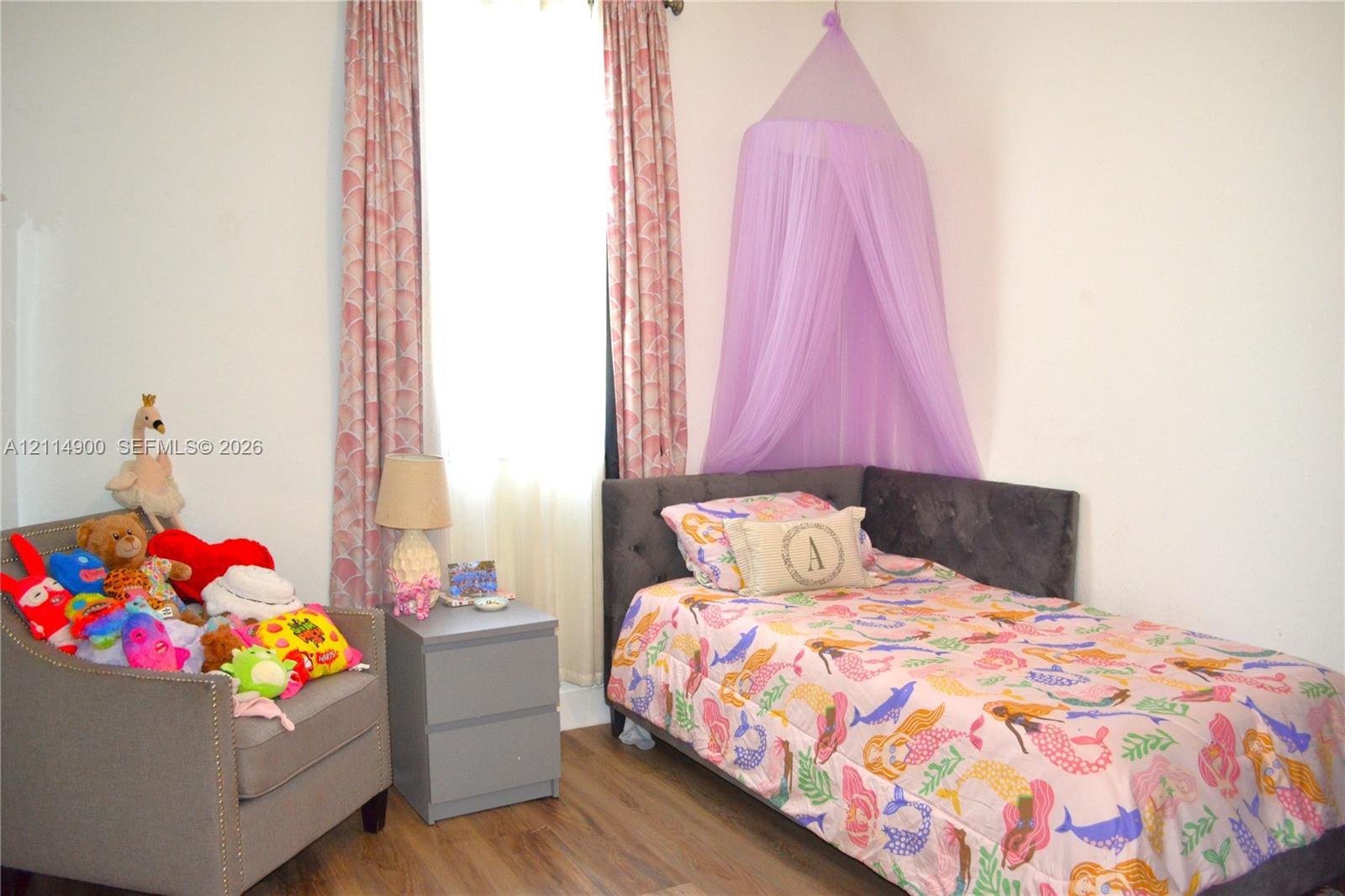 Neutral-toned interior space featuring wood-finish flooring and a single vertical window dressed with floor-length patterned curtains