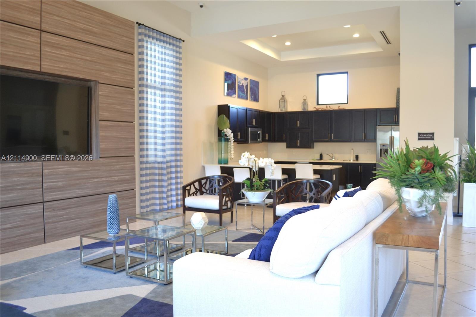 Large-scale wood-finished media wall, recessed tray ceiling, kitchen cabinetry with dark finish, and tile flooring