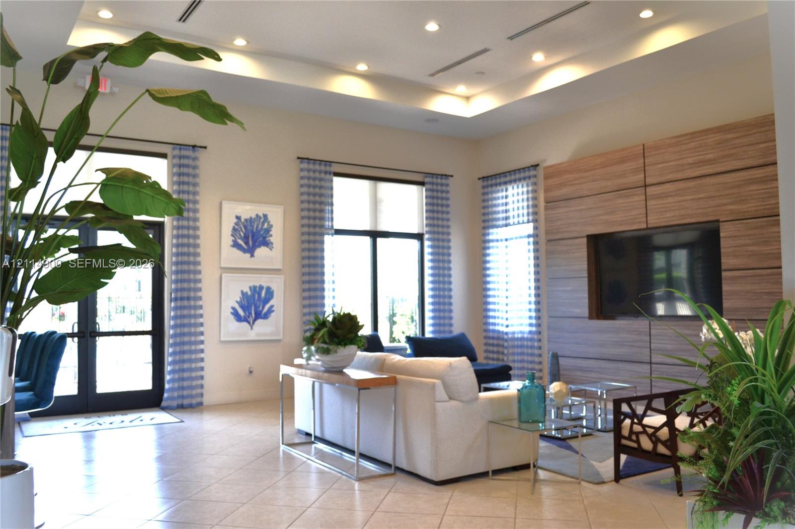Open-concept living space featuring high tray ceilings with recessed lighting, large-format tile flooring, oversized windows, and a wood-panel accent wall with integrated media storage