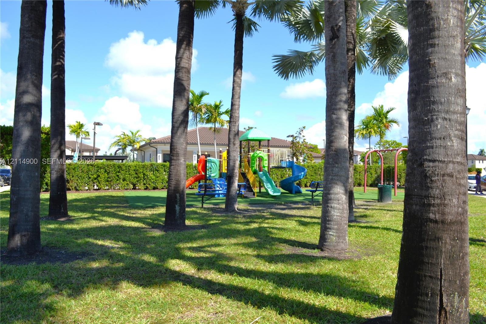 Outdoor recreational area featuring mature palm trees, manicured lawn, hedge landscaping, and a multi-component play structure