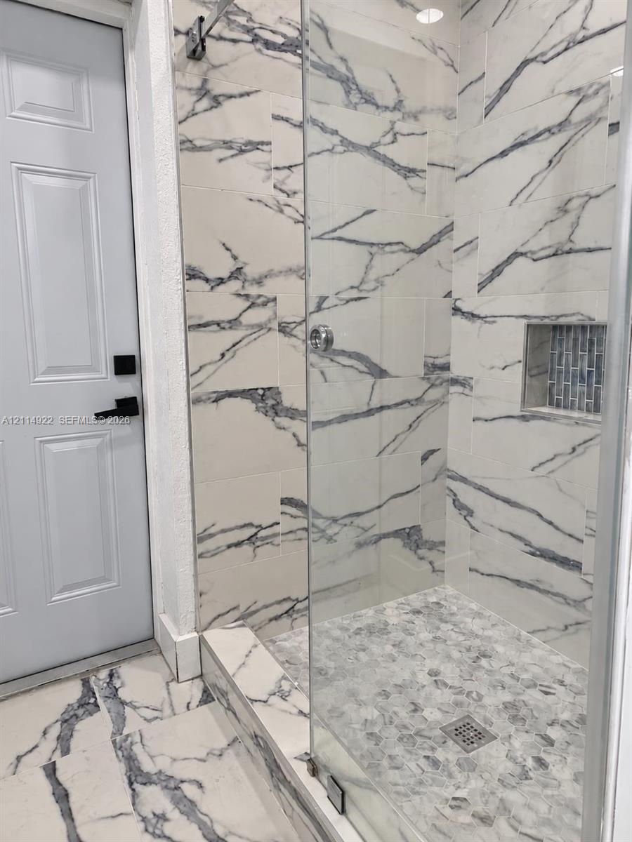 Walk-in shower featuring large-format veined tile surround, glass enclosure, pebble-stone flooring, built-in recessed niche with mosaic accent tile, and matte black door hardware