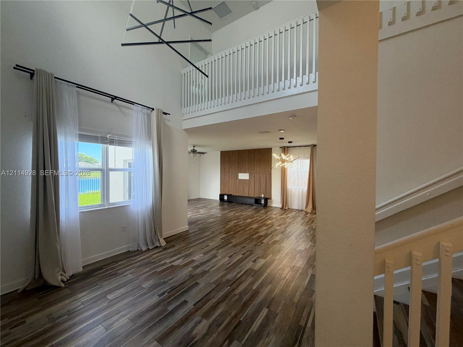 Double-height ceiling with geometric chandelier, wood-finish flooring, second-level balcony railing, and wood-paneled accent wall