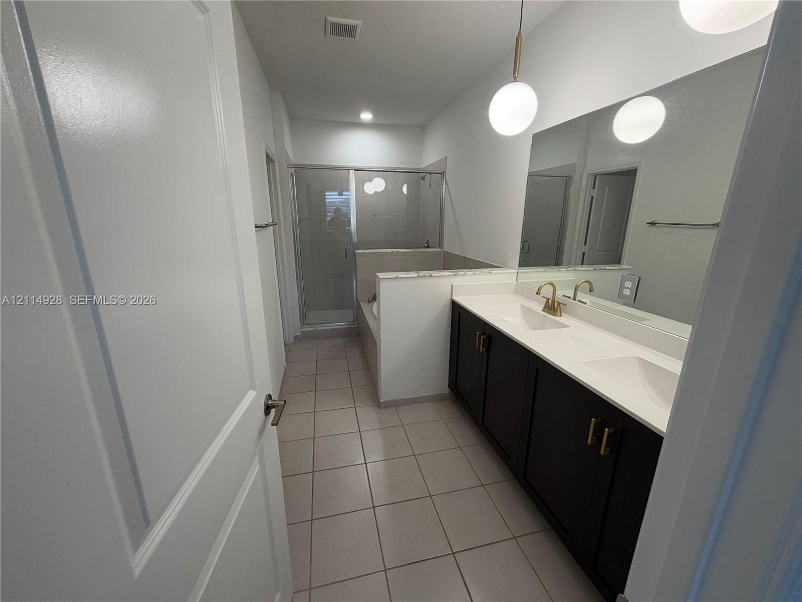 Dual-sink vanity with dark cabinetry and white countertops, pendant globe lighting, large wall-mounted mirror, ceramic tile flooring, and a glass-enclosed shower with a soaking tub