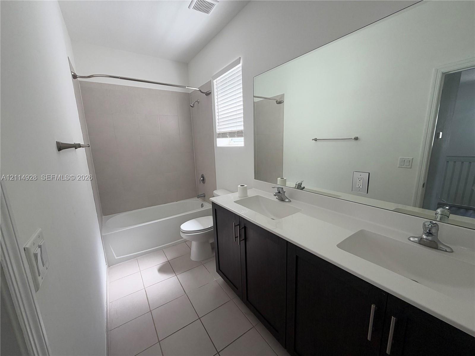 Bathroom featuring a dark-finish dual-sink vanity with white countertops, large wall-mounted mirror, ceramic tile flooring, and a bathtub with tile surround