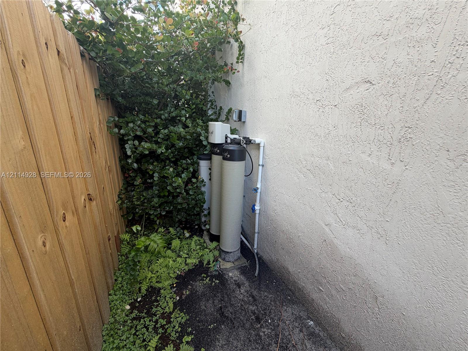 Water filtration system mounted to the exterior wall, enclosed by a wood-plank fence and mature landscaping