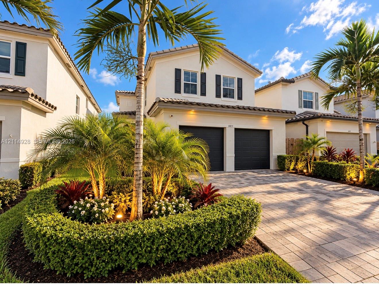 Two-story stucco exterior featuring a paver driveway, two-car garage, tile roofing, and mature palm tree landscaping