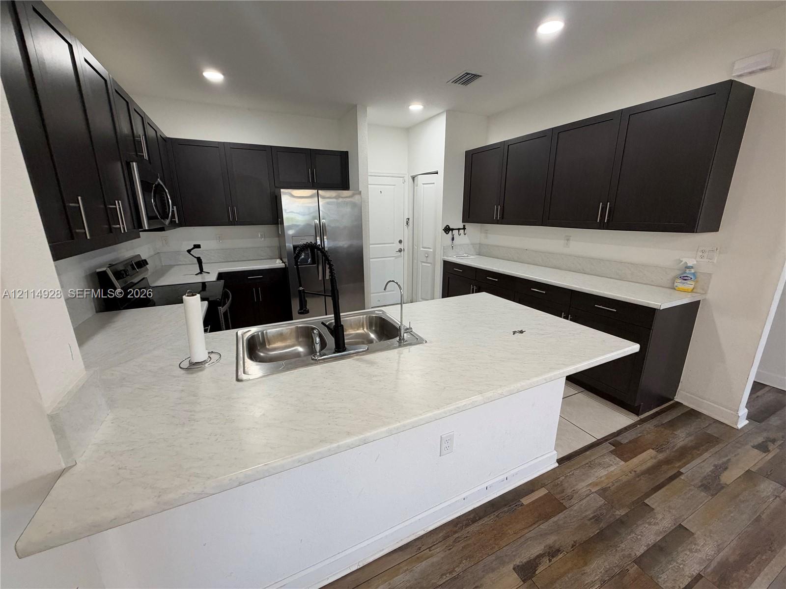 Kitchen featuring dark wood cabinetry, a central island with a dual-basin sink, light-toned countertops, stainless steel appliances, and wood-finish flooring