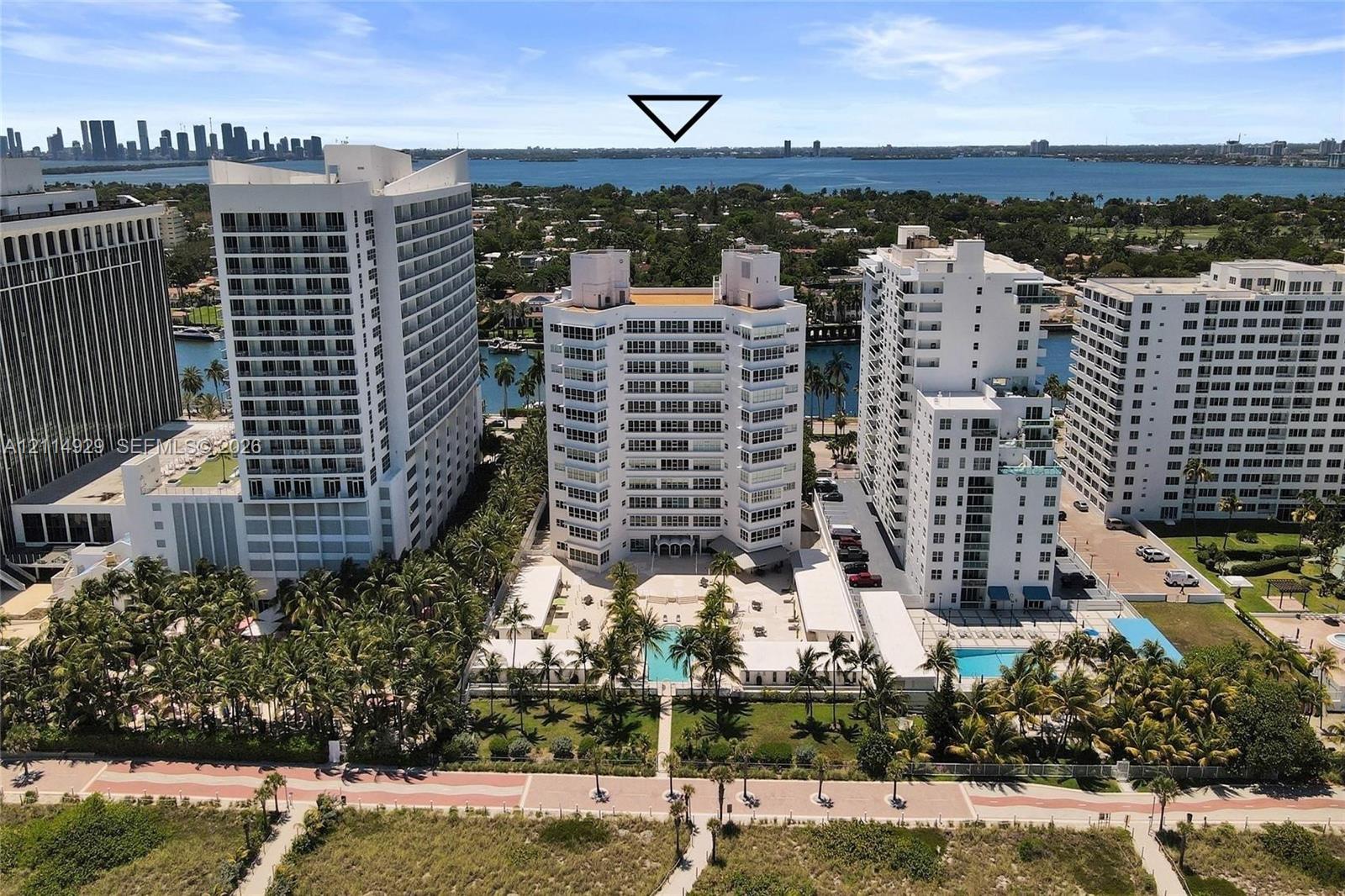 Aerial perspective of a high-rise residential complex featuring a swimming pool, paved hardscaping, mature palm trees, and coastal water surroundings