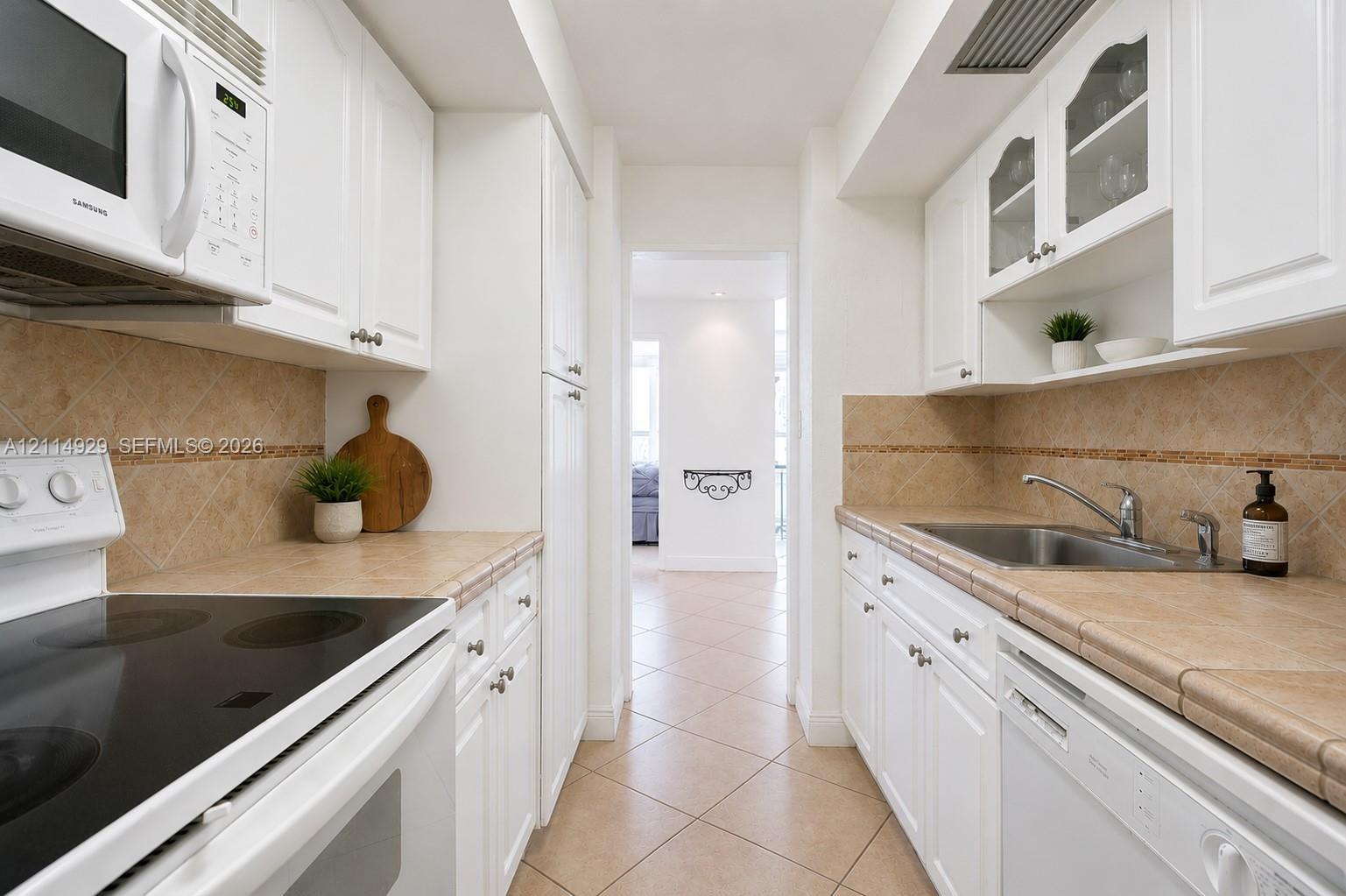 Galley kitchen featuring white cabinetry, tile countertops with matching backsplash, under-cabinet lighting, and glass-front upper cabinet doors
