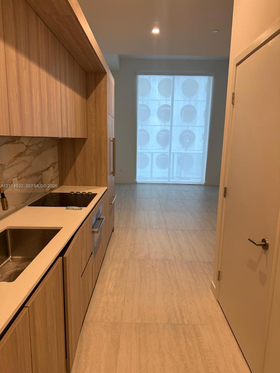 Kitchen featuring wood-finish cabinetry, stone-surface countertops, full-height decorative backsplash, stainless steel sink, and large-format tile flooring