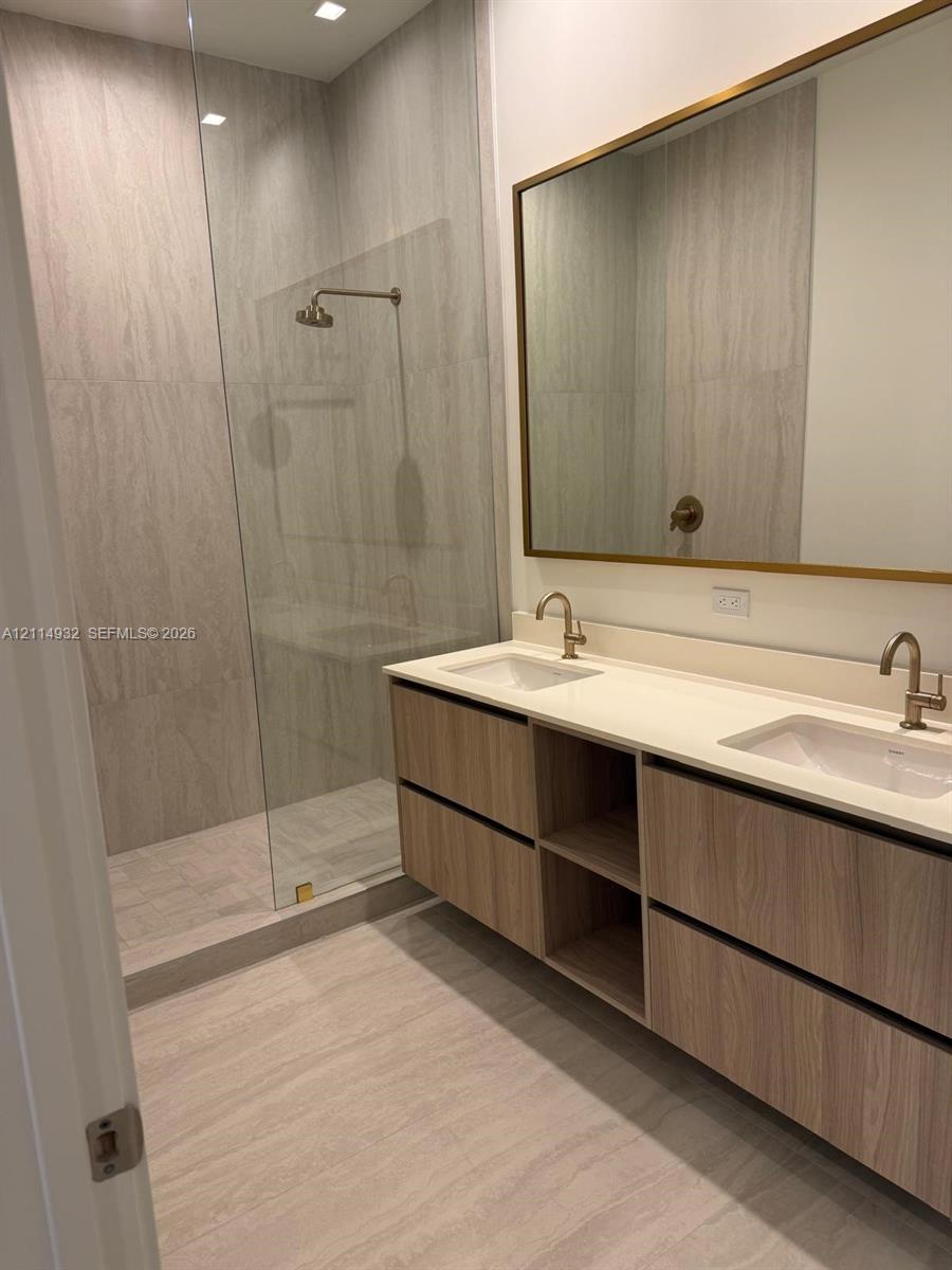Floating dual-vanity with wood-finish cabinetry, white countertop, integrated rectangular sinks, brass-toned fixtures, and a glass-enclosed shower with floor-to-ceiling tile