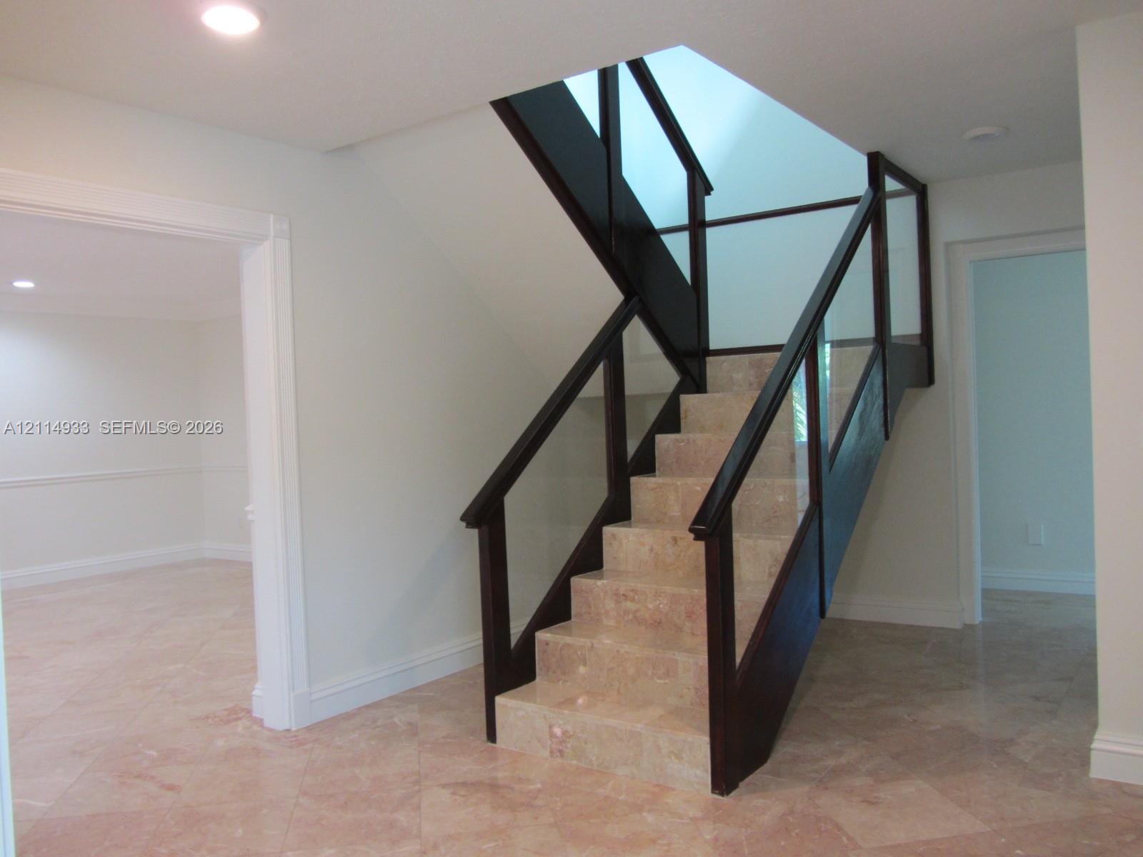 Staircase featuring natural stone treads, dark wood handrails, and glass panel balustrades beneath a skylight Staircase featuring natural stone treads, dark wood handrails, and glass panel balustrades beneath a skylight