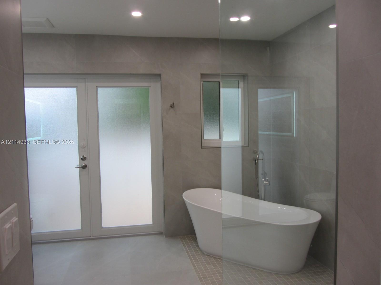 Freestanding soaking tub with a floor-mounted faucet, large-format grey tile wall cladding, glass shower enclosure, and white frosted glass double doors